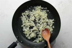 Diced white onions being sautéed in a black non-stick frying pan with a wooden spoon on a white countertop.