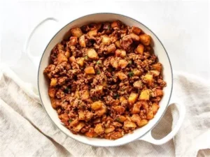 A top-down view of a white ceramic skillet filled with cooked ground turkey, roasted potato cubes, and fresh herbs on a light linen background.