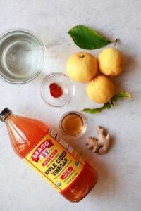 Fresh ingredients for an apple cider vinegar drink recipe for gut health, including a bottle of Bragg ACV, raw ginger, lemons, and honey.
