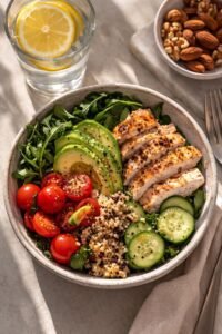 A nourishing meal bowl filled with sliced grilled chicken breast, avocado, quinoa, cherry tomatoes, cucumbers, and fresh greens, served alongside a glass of lemon water and a small bowl of mixed nuts.