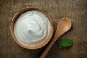 Top-down view of thick, creamy Greek yogurt in a wooden bowl with a wooden spoon and a mint leaf on a rustic surface.