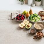 Array of high-fiber foods including berries, avocado, apples, legumes, seeds, and whole-grain bread on a light wooden table.
