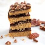A tall stack of homemade Low Carb Desserts featuring shortbread-style bars topped with dark chocolate fudge and chopped pecans on a light surface.