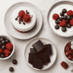 A flat-lay selection of low carb sweet snacks including mini berry cheesecake, dark chocolate squares, Greek yogurt with berries, mixed nuts, and fresh blueberries.